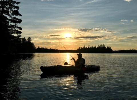 The silhouette of a person in a kayak holding a fishing rod while floating on a large body of water. The sun sets behind them. The sky is colored with vibrant blues and yellows.