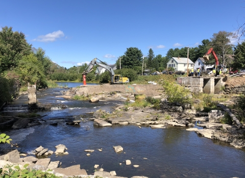 Concrete dam spans halfway across a river with heavy machinery and people in construction hats on top