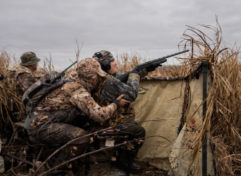 Person is camouflage hunting gear crouches behind a second person in a wheelchair in a duck hunting blind
