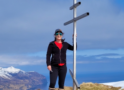 A woman wearing a hat and sunglasses on a high summit overlooking snowcapped mountains and ocean. She stands next to a metal structure and a buoy that sits on the ground.