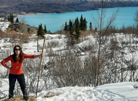 A woman standing wearing a bright orange shirt and sunglasses holding a walking stick in a snowy landscape. A bright, aqua blue lake in the background.