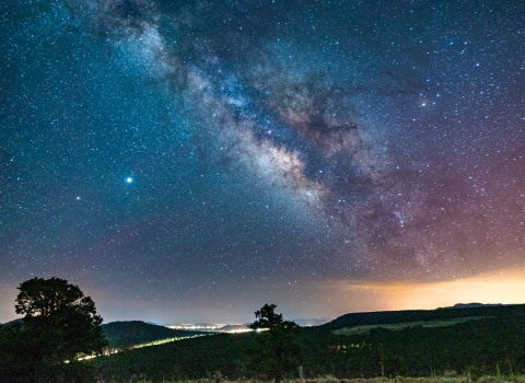 A bright light obstructs the night sky and milkyway directly over a city at night with hills and trees in the foreground and stars pictured in the background.