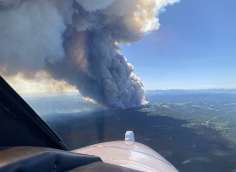 A view of a smoke plume from an airplane. The plume of smoke is billowing into blue sky from a forested landscape.