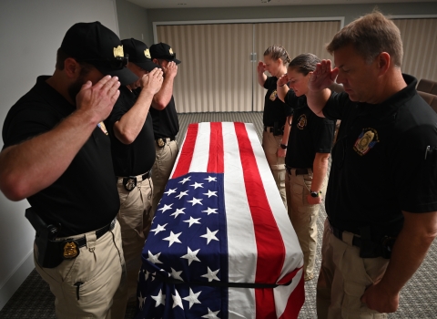 The honor guard stands over a coffin draped in an American flag. Each of them is saluting and looking down at the coffin/flag.