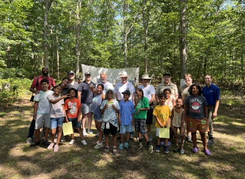a large group of kids and adults smile in front of an archery range in the forest