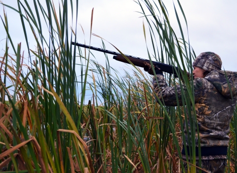 Female standing in tall grass wearing camouflage hunting outfit holding a rifle and aiming to the left.
