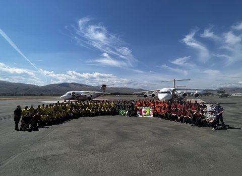 A group of people stand together in a semi circle on a tarmac in front of aircraft used for firefighting efforts. The group has flags out and are all in firefighter clothing or agency t-shirts.