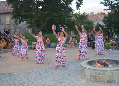  7 people in similar dresses with hands raised to left in unison dance outdoor amphitheater 