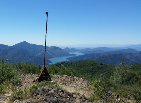 A tall pole on the top of a mountain with a microphone at the top. This pole is secured with anchors to the rocks below. In the background is a bright blue river flowing through the valley of mountains. 
