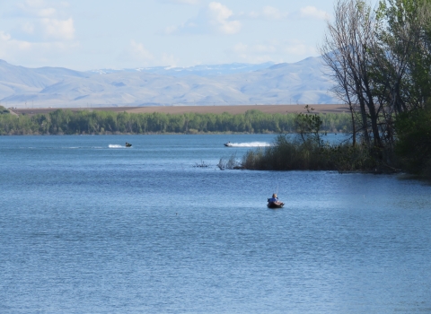 A kayaker is fishing and two people are riding personal watercraft on a calm lake. There are trees on a point in the foreground and on the distant shoreline, as well as mountains in the distance. 
