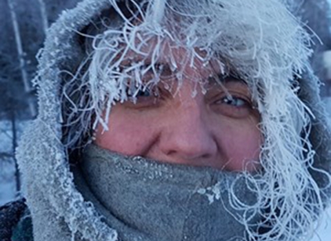 A person with frost-covered hair peeking out of a hood. Their mouth is covered by a neck gaiter and they smile with their eyes.