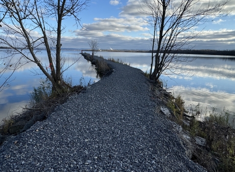 a stone jetty with sparse trees cuts through a still lake that reflects blue skies and clouds