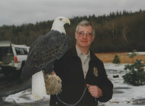 A bald eagle perches on the arm of a man in a Service jacket in a snowy forested outdoor setting