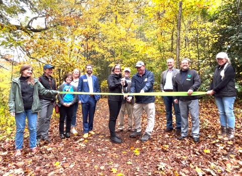 A group of people stand in front of a forest holding a ribbon while a woman cuts the ribbon with a huge pair of scissors. 