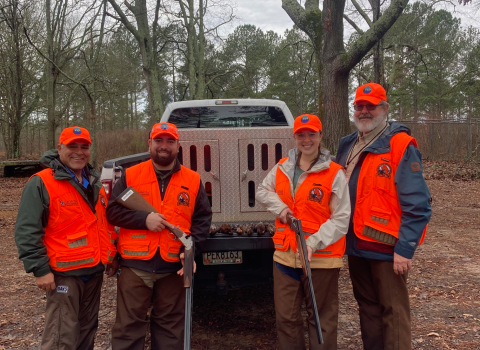 Four people in orange hunting vests, two with firearms in their hands, stand in front of a truck in the woods smiling