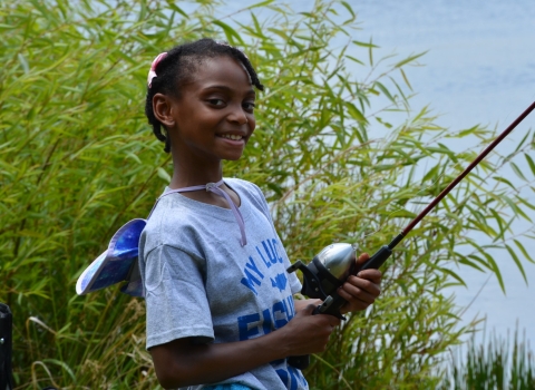 Photo of smiling girl about 10 years old holding a fishing rod with willows and a pond in the background.