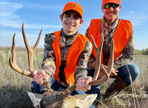 Two young brothers wearing orange and camo with the younger one holding a harvest mule deer by the antlers