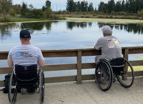 Two men in manual wheelchairs sit side by side fishing from a paved path, looking out over a calm pond bordered by trees and greenery, with a wooden railing in front of them.