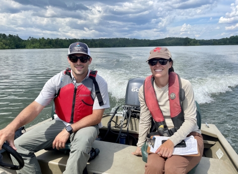 Two people sit on a moving boat, with one person driving while the other holds a clipboard