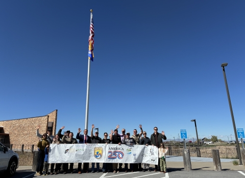 Group of people standing outdoors holding a USFWS-America 250 banner beneath a flagpole; flags flat