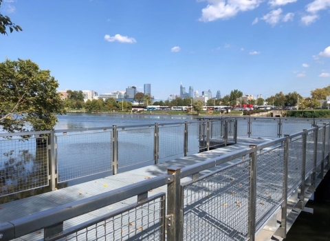 View of a pier with metal railings and synthetic decking jutting out into a river, with a city skyline in the distance