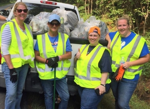 Four women in bright safety vests stand in front of a white pickup truck full of bags of roadside litter.