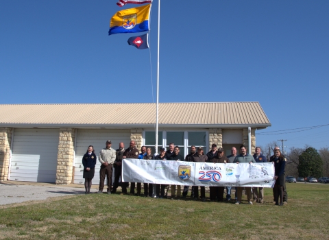 People hold FWS A250 banner at base of flagpole with US flag, FWS flag, fish pennant flying