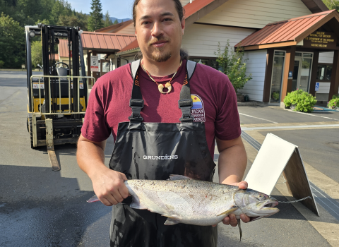 Addison Emerson holding an adult coho salmon at Quilcene National Fish Hatchery. Forklift, facility, and skyline in the background.