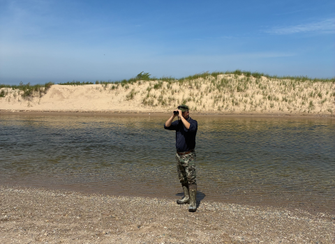 A man standing on a beach looking through binoculars away from the water.