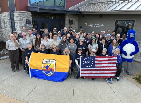Large group posing outside a building with sign on it saying “JOHN D. DINGELL JR. VISITOR CENTER.” They’re holding USFWS flag and Freedom 250 U.S. flag; blue mascot stands at right