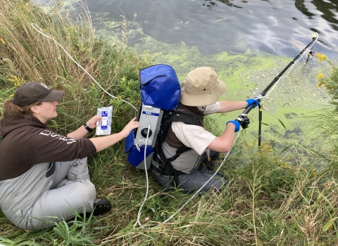 Two USFWS biologists use an eDNA backpack sampler to collect water samplers from the shore.