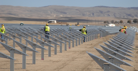rows of gray solar panels stand out against the dry desert background. Solar panel installers wearing yellow vests work on the panels. 